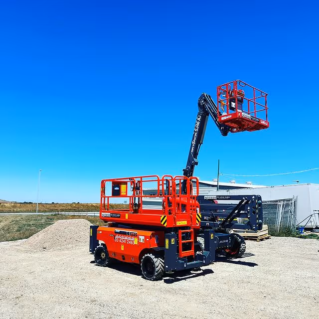 A red cherry picker with an extended arm stands on gravel under a clear blue sky.