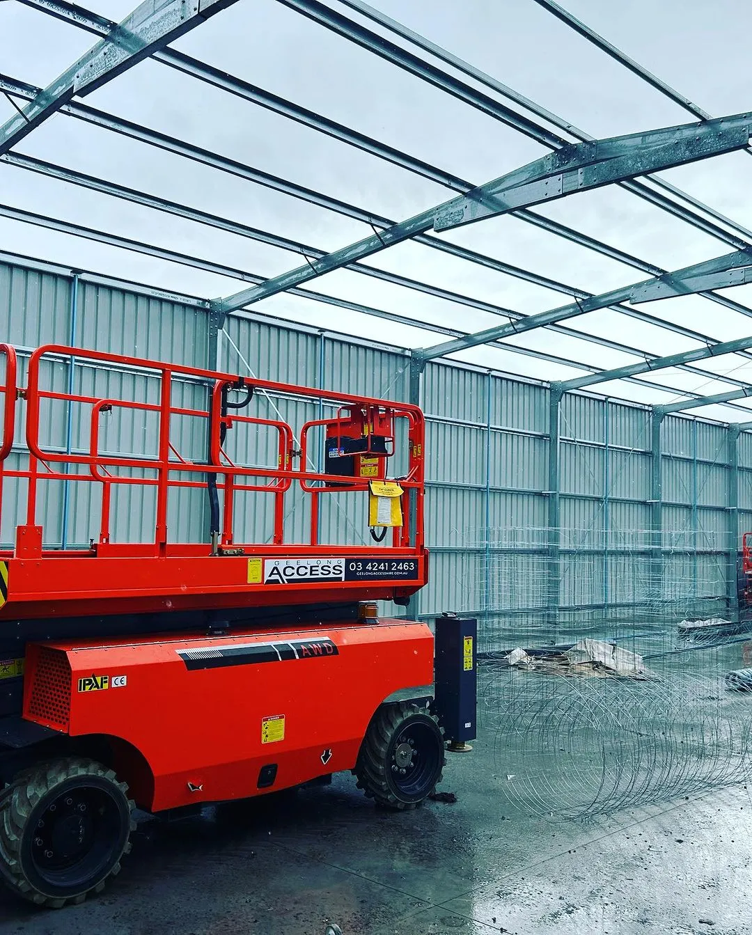 A red scissor lift is inside a large, unfinished metal-framed building under construction.